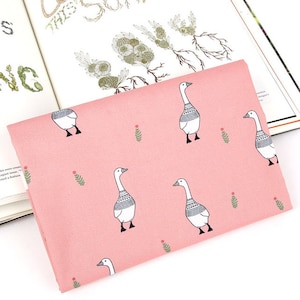 May include: Pink fabric with a repeating pattern of white geese with gray and black accents. The geese are interspersed with small green sprigs and red dots. The fabric is folded and lies on top of an open book.