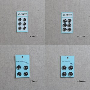 May include: Four packs of black snap fasteners in different sizes: 12mm, 14mm, 17mm, and 19mm. Each pack has a blue label with the text "Snap Fasteners" and the size of the fasteners printed on it.