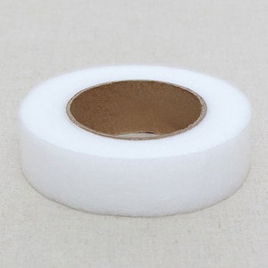 May include: A roll of white, non-woven fabric with a brown cardboard core. The fabric is tightly wound, creating a circular shape. The image is a close-up, showing the texture of the fabric.