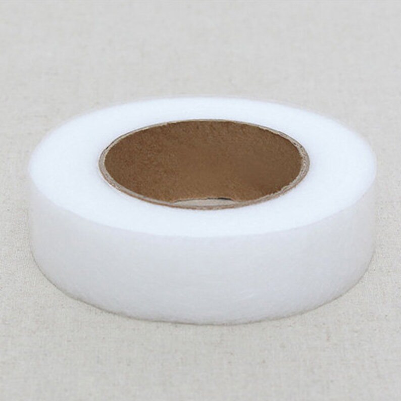 May include: A roll of white, non-woven fabric with a brown cardboard core. The fabric is tightly wound, creating a circular shape. The image is a close-up, showing the texture of the fabric.