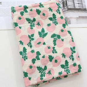 May include: A folded piece of fabric featuring an off-white background with a repeating pattern of light pink tomatoes and vibrant green leaves and stems. The textile has a soft, textured appearance with a consistent print across the surface.