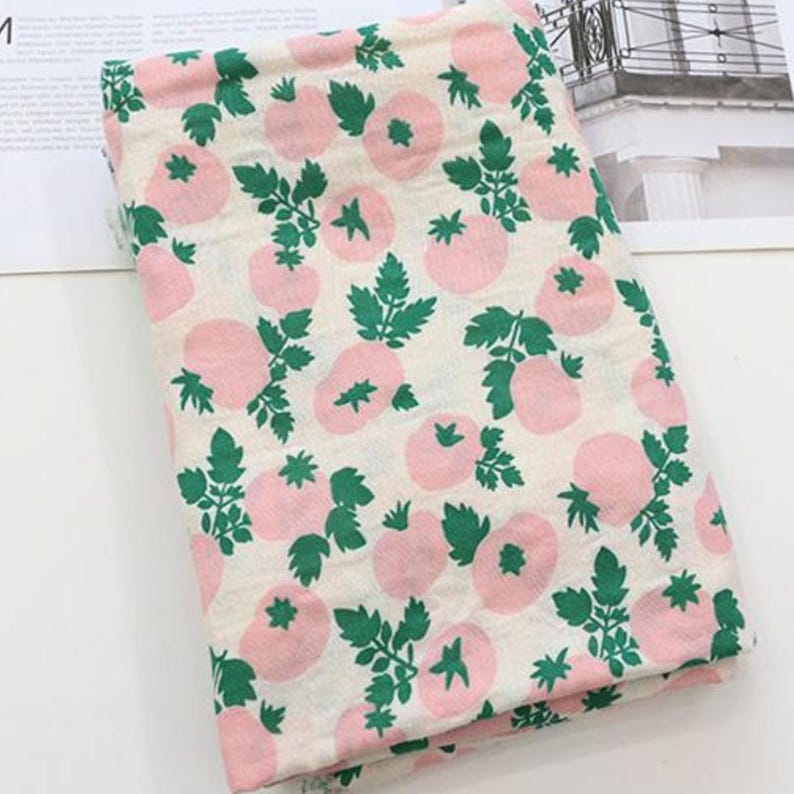 May include: A folded piece of fabric featuring an off-white background with a repeating pattern of light pink tomatoes and vibrant green leaves and stems. The textile has a soft, textured appearance with a consistent print across the surface.