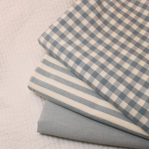 May include: Three folded pieces of fabric stacked together. The top piece features a blue and white gingham check pattern. The middle piece displays blue and white vertical stripes. The bottom piece is a solid, plain light blue fabric.