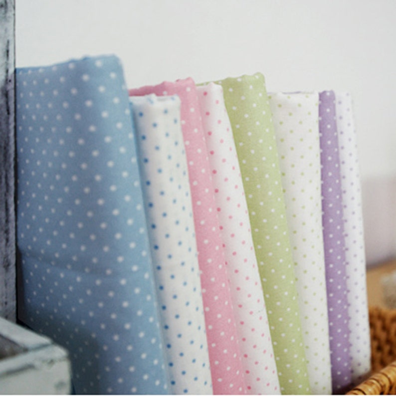 May include: A collection of folded fabric bolts arranged in a row, featuring a repeating white polka dot pattern on various pastel colors including light blue, pink, lime green, white, and lavender, showcasing a soft, woven textile texture.