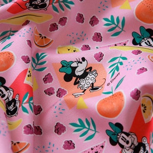 Puede incluir: Tela rosa con un patr&oacute;n repetido de Minnie Mouse, fruta y hojas. La tela presenta un fondo rosa con lunares blancos. El patr&oacute;n incluye a Minnie Mouse en varias poses, rodajas de sand&iacute;a, naranjas, limones, frambuesas y hojas verdes. El texto "&copy; Disney" es visible en la esquina inferior derecha.