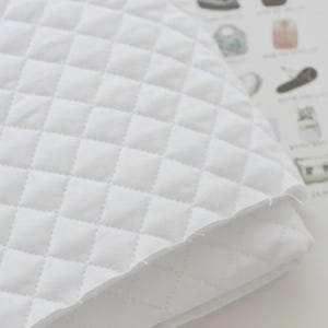 May include: A white quilted fabric featuring a repeating diamond-shaped stitched pattern. The material has a soft, layered appearance with visible raw edges along the folded corner, showcasing the texture and construction of the textile.