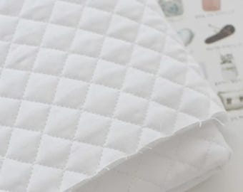 May include: A white quilted fabric featuring a repeating diamond-shaped stitched pattern. The material has a soft, layered appearance with visible raw edges along the folded corner, showcasing the texture and construction of the textile.