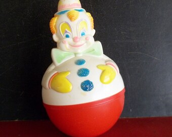 May include: A vintage, white plastic toy clown with a red and white hat, blue eyes, and a red nose. The clown is wearing a green bow tie and has a white body with blue and white polka dots. The clown is a rocking toy with a red base.