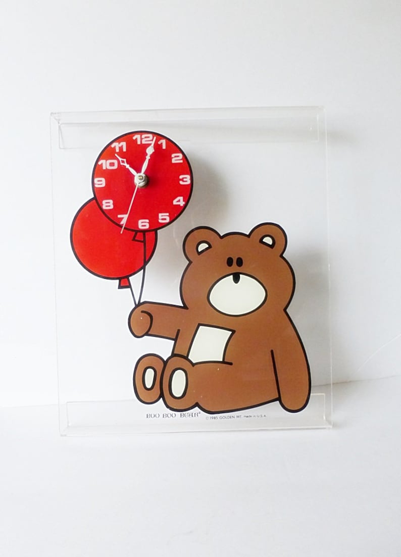 May include: A clear acrylic clock with a brown bear holding two red balloons. The clock face is red with white numbers and hands. The clock reads "BOO BOO BEAR" and "1985 GOLDEN MESA, INC. U.S.A."