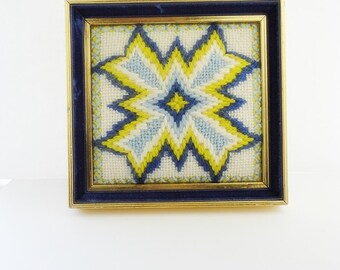 May include: A framed needlepoint piece with a yellow, blue, and white geometric star design.
