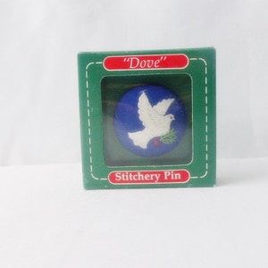 May include: A white dove with an olive branch in its beak is embroidered on a blue circle. The circle is surrounded by a green border with the text "Dove" and "Stitchery Pin".