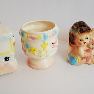 May include: Three vintage ceramic planters, one shaped like a train, one shaped like a girl holding a teddy bear, and one shaped like a flower pot with a blue and yellow ribbon design. The girl planter has the word "GIRL" spelled out in blocks.