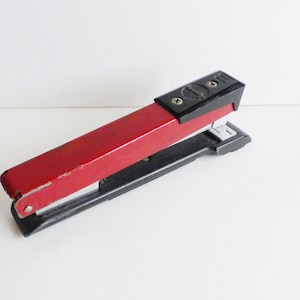 May include: A red and black stapler with a silver staple remover. The stapler has a black plastic top and a red metal body.
