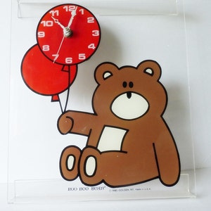 May include: A brown teddy bear holding two red balloons with a clock face on the top balloon. The clock face has white numbers and hands. The text "BOO BOO BEAR" and "1985 GOLDEN MT made in USA." is printed below the bear.