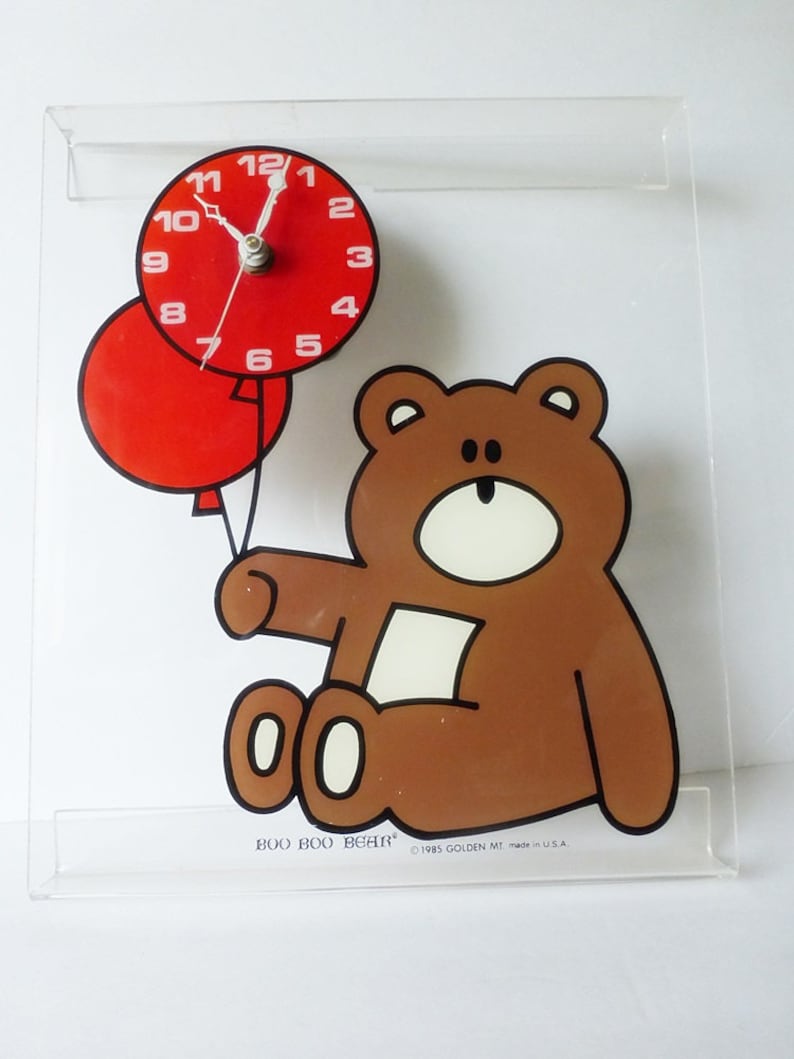 May include: A brown teddy bear holding two red balloons with a clock face on the top balloon. The clock face has white numbers and hands. The text "BOO BOO BEAR" and "1985 GOLDEN MT made in USA." is printed below the bear.