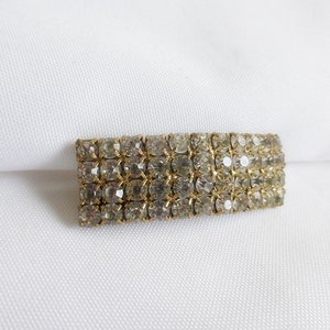 Puede incluir: Un broche de barra de tono dorado con forma rectangular, cubierto de diamantes de imitación transparentes.