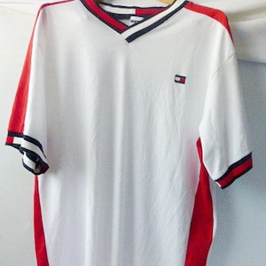 May include: White short-sleeved t-shirt with red and blue trim on the collar and sleeves. The shirt has a red and white side panel and a small Tommy Hilfiger logo on the chest.