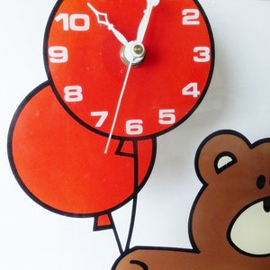 May include: A red clock with black hands and white numbers is attached to a red balloon. A brown teddy bear is holding the balloon string. The clock face reads 12:01.