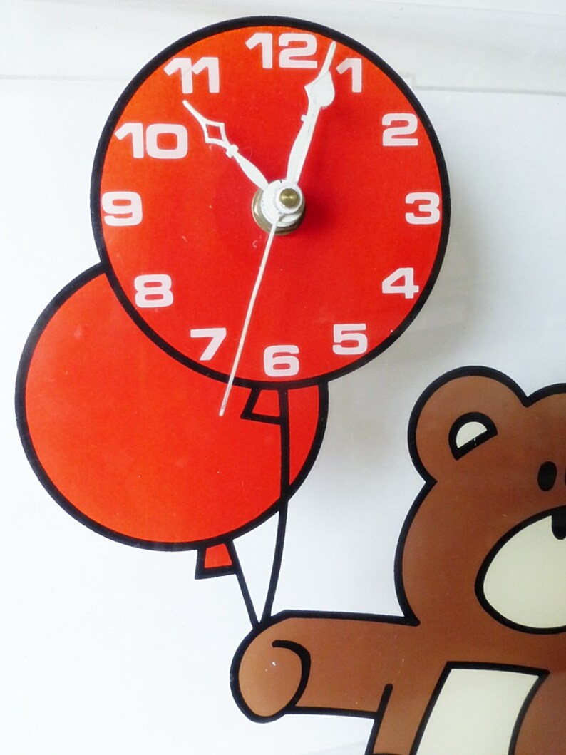 May include: A red clock with black hands and white numbers is attached to a red balloon. A brown teddy bear is holding the balloon string. The clock face reads 12:01.