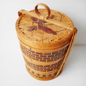 May include: A round, woven bamboo basket with a handle. The basket has a decorative design on the lid and is made of natural brown and dark brown bamboo.