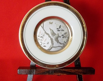 Könnte beinhalten: Ein weißer Porzellanteller mit goldfarbenem Rand und einem silberfarbenen Design von zwei Vögeln auf einem Ast mit einem Schmetterling. Der Teller wird auf einem Holzständer präsentiert.