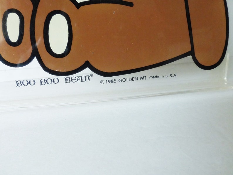 May include: A clear plastic Boo Boo Bear lunchbox with a brown bear cartoon illustration. The lunchbox has the text "Boo Boo Bear" and "&copy; 1985 GOLDEN MT. made in U.S.A."