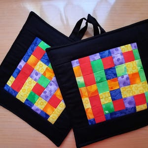 Könnte beinhalten: Zwei quadratische, schwarze Topflappen mit farbenfrohen Patchwork-Mittelstücken. Das Patchwork zeigt ein Gitter aus roten, orangefarbenen, gelben, grünen, blauen und violetten Quadraten. Jeder Topflappen hat eine schwarze Schlaufe zum Aufhängen.
