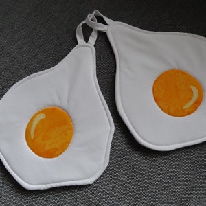 Könnte beinhalten: Zwei weiße, eiförmige Topflappen mit gelbem Eigelb-Design. Jeder Topflappen hat eine Schlaufe zum Aufhängen. Die Topflappen sind aus einem weichen, gesteppten Material gefertigt und sollen Spiegeleiern ähneln.