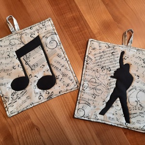 Könnte beinhalten: Zwei quadratische Topflappen mit Musikthema. Einer zeigt eine große schwarze Musiknote, der andere eine schwarze Silhouette eines Interpreten. Beide haben einen cremefarbenen Hintergrund mit Noten und eine Aufhängeschlaufe.