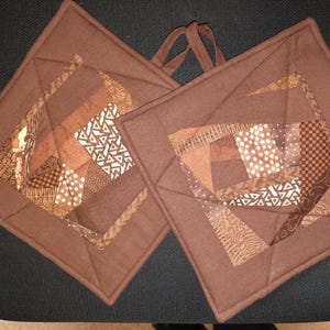 Könnte beinhalten: Zwei braune, gesteppte Topflappen mit Patchwork-Design. Jeder quadratische Topflappen hat ein zentrales geometrisches Muster in verschiedenen Braun- und Beigetönen mit einer Schlaufe zum Aufhängen.