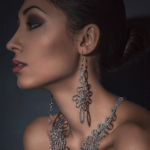 May include: A silver-coloured necklace and matching earrings set. The jewellery features an intricate, openwork design. The earrings hang down from the ear, and the necklace drapes around the neck. The background is a dark, solid colour.