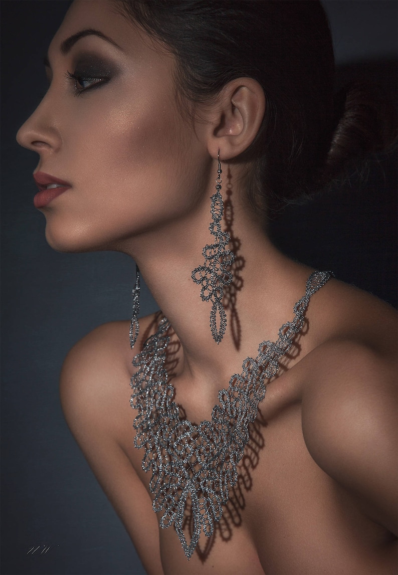 May include: A silver-coloured necklace and matching earrings set. The jewellery features an intricate, openwork design. The earrings hang down from the ear, and the necklace drapes around the neck. The background is a dark, solid colour.