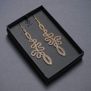 May include: Gold-coloured, ornate dangle earrings presented in a black box. The earrings have an intricate, openwork design with a floral and geometric pattern. The box is on a dark grey surface.