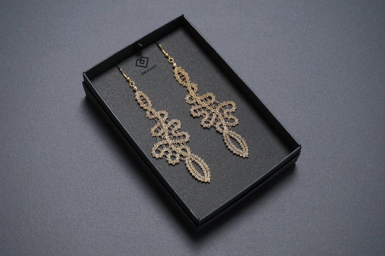 May include: Gold-coloured, ornate dangle earrings presented in a black box. The earrings have an intricate, openwork design with a floral and geometric pattern. The box is on a dark grey surface.