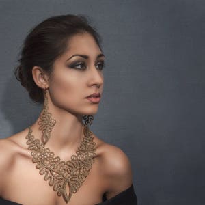 May include: A woman wearing a gold-coloured lace necklace and matching earrings. The intricate jewellery is the focal point, set against a dark background. The woman is wearing a black off-the-shoulder top.