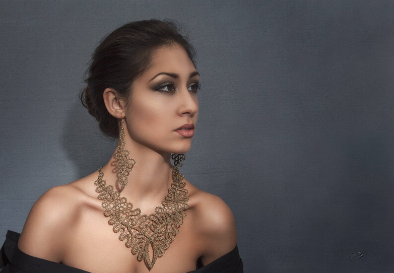 May include: A woman wearing a gold-coloured lace necklace and matching earrings. The intricate jewellery is the focal point, set against a dark background. The woman is wearing a black off-the-shoulder top.