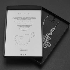 May include: A monochrome image showing a jewellery box with a pair of lace earrings. The box is open, displaying a printed card with text about lace jewellery and a map of Slovenia. The earrings are detailed and intricate.