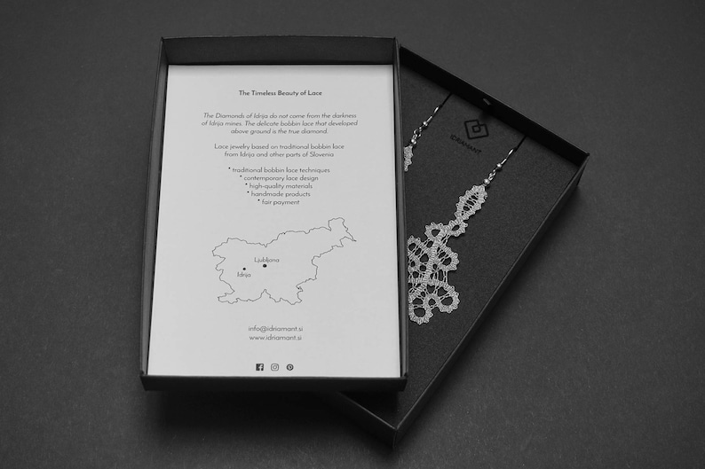 May include: A monochrome image showing a jewellery box with a pair of lace earrings. The box is open, displaying a printed card with text about lace jewellery and a map of Slovenia. The earrings are detailed and intricate.