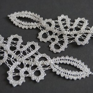 May include: Two pieces of silver lace trim with an openwork design. The lace has a delicate, intricate pattern with scalloped edges. The trim is displayed on a dark grey background.