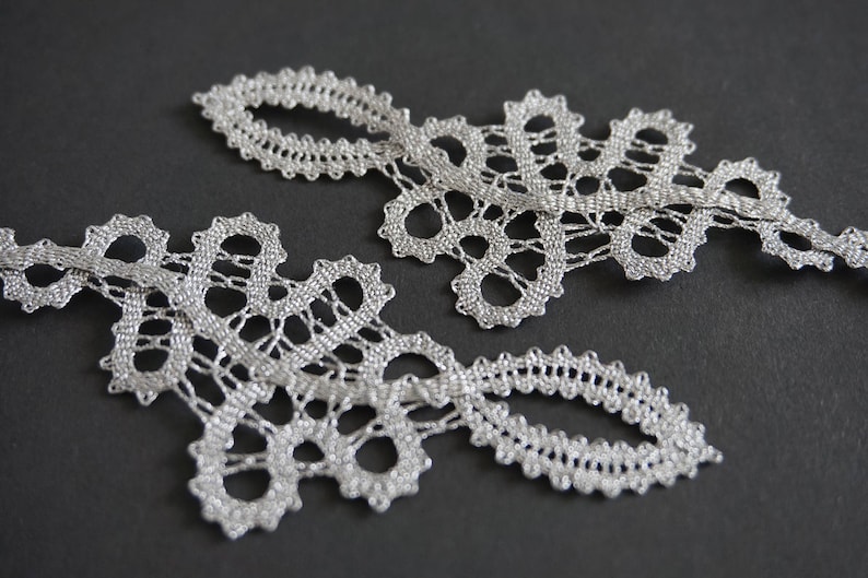 May include: Two pieces of silver lace trim with an openwork design. The lace has a delicate, intricate pattern with scalloped edges. The trim is displayed on a dark grey background.