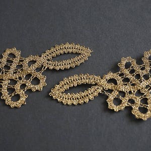 May include: Close-up of two sections of gold lace trim. The lace features a delicate, openwork design with floral-like shapes and oval accents. The trim is set against a dark grey background, highlighting the intricate details.
