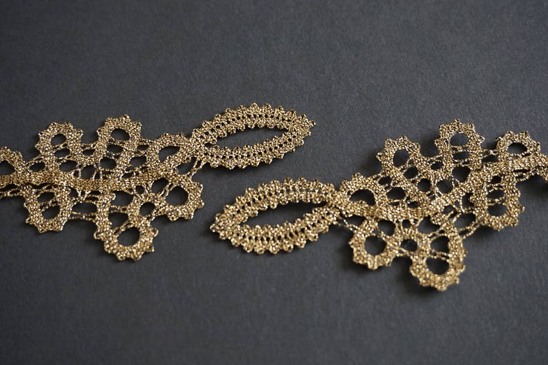 May include: Close-up of two sections of gold lace trim. The lace features a delicate, openwork design with floral-like shapes and oval accents. The trim is set against a dark grey background, highlighting the intricate details.