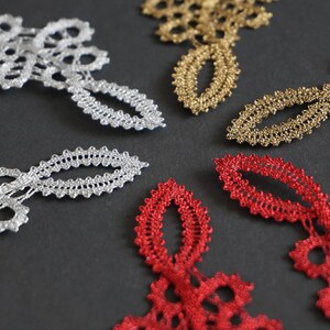 May include: Close-up of decorative lace pieces in silver, gold, and red. The lace features a delicate, openwork design with floral and oval shapes. The pieces are arranged on a dark grey surface.