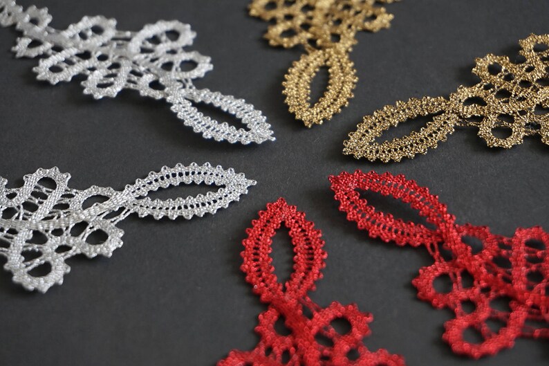 May include: Close-up of decorative lace pieces in silver, gold, and red. The lace features a delicate, openwork design with floral and oval shapes. The pieces are arranged on a dark grey surface.