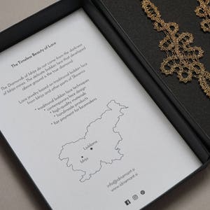 May include: Gold-coloured lace earrings presented in a black box. The box includes a printed insert with text about the earrings and a map of Slovenia. The earrings feature an intricate design.
