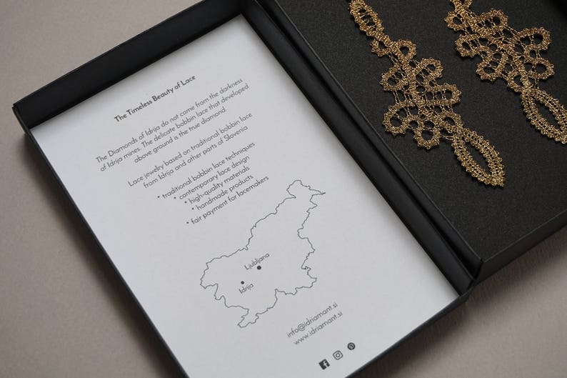 May include: Gold-coloured lace earrings presented in a black box. The box includes a printed insert with text about the earrings and a map of Slovenia. The earrings feature an intricate design.