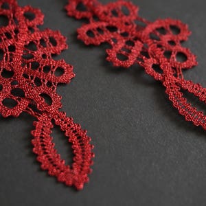 May include: Close-up of two intricate, red lace ornaments against a dark grey background. The lace features a detailed, openwork design with a teardrop-shaped bottom. The ornaments appear delicate and handmade.