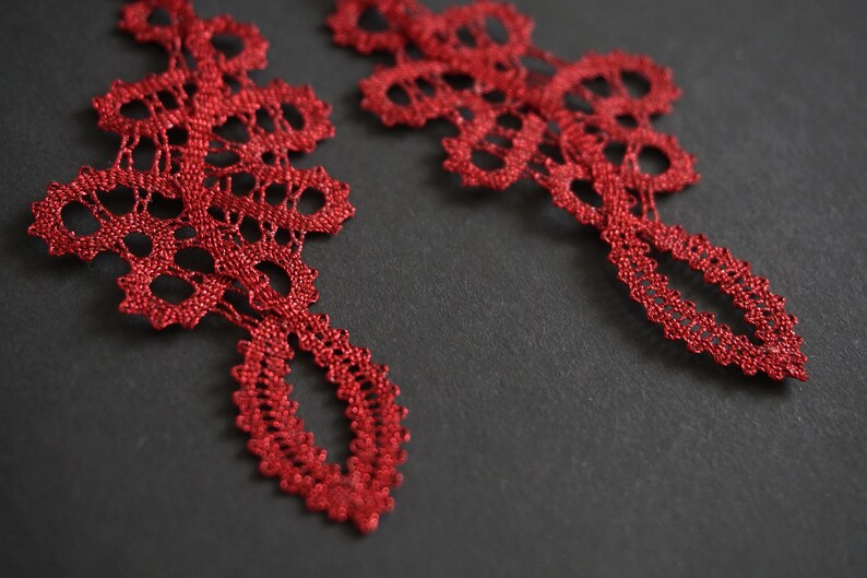 May include: Close-up of two intricate, red lace ornaments against a dark grey background. The lace features a detailed, openwork design with a teardrop-shaped bottom. The ornaments appear delicate and handmade.
