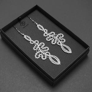 May include: A pair of white lace earrings presented in a black box. The earrings feature a delicate, openwork design with intricate floral and geometric patterns. The earrings are suspended from silver-coloured hooks.