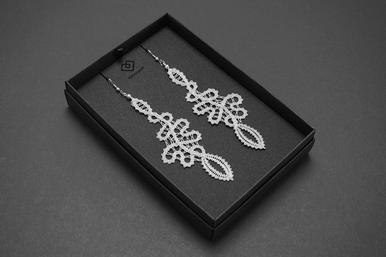 May include: A pair of white lace earrings presented in a black box. The earrings feature a delicate, openwork design with intricate floral and geometric patterns. The earrings are suspended from silver-coloured hooks.